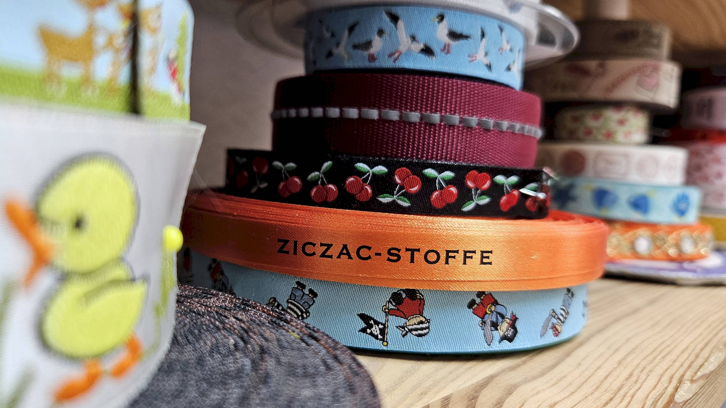 Nahaufnahme verschiedener aufgerollter Bänder und Borten mit unterschiedlichen Farben und Mustern. Im Mittelpunkt liegt ein orangefarbenes Satinband mit der Aufschrift „ZICZAC-STOFFE“. Darum sind unter anderem hellblaue Bänder mit Figuren und Vögeln, ein schwarzes Band mit roten Kirschen sowie weitere gemusterte Stoffbänder angeordnet. 